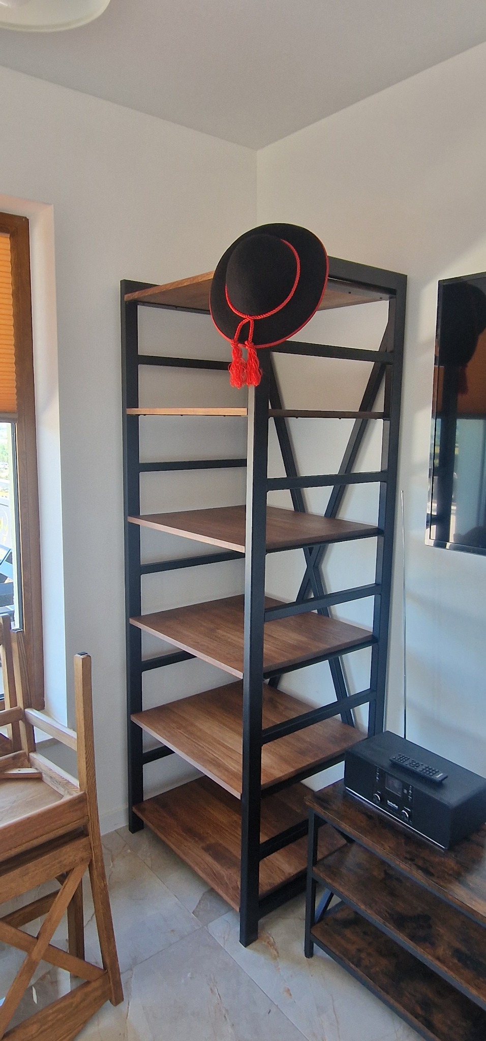 Regał metalowy z drewnianymi półkami w stylu industrialnym, ozdobiony czarnym kapeluszem z czerwonymi pomponami. Widok z boku, jasne wnętrze, fragment krzesła.
