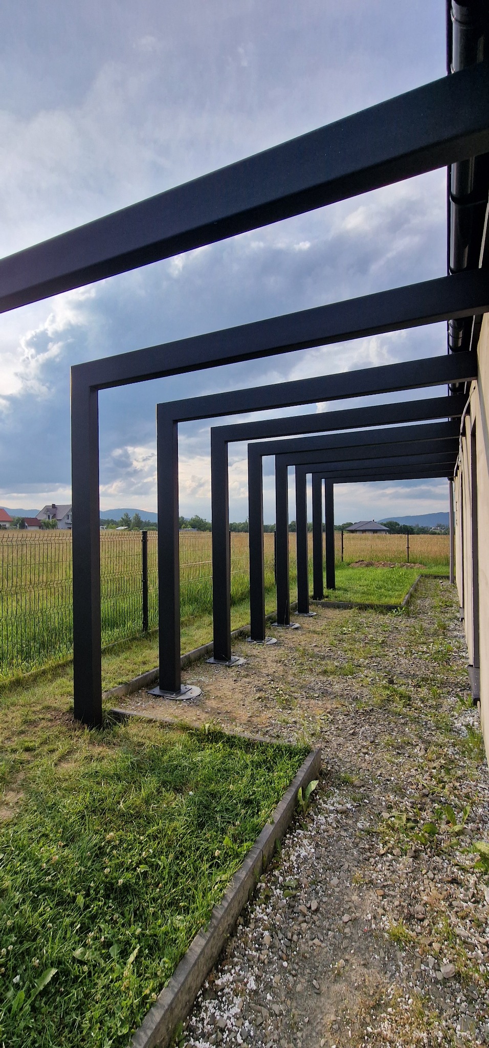 Nowoczesna pergola metalowa w kolorze czarnym, z widokiem na zielony krajobraz i błękitne niebo z chmurami. Minimalistyczny design.