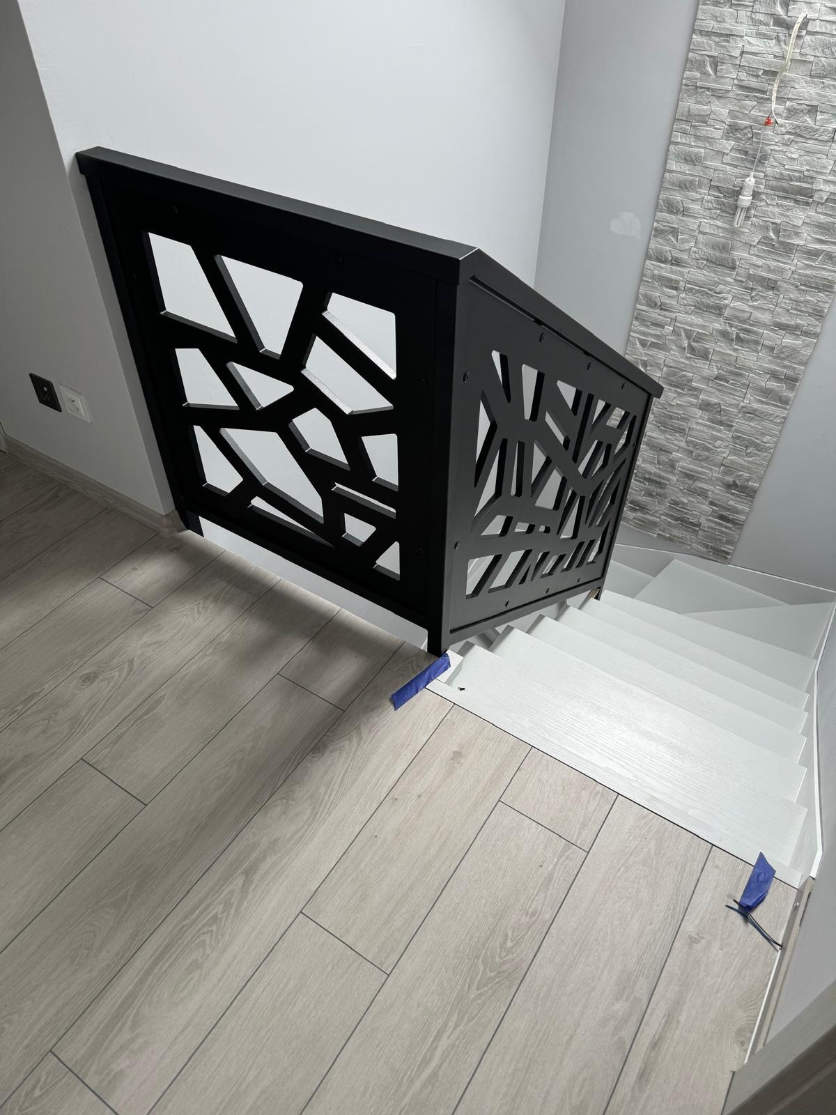 Nowoczesna, czarna balustrada schodowa z geometrycznym wzorem, zamontowana na białych schodach. Ściana z dekoracyjną okładziną w tle. Parkiet w odcieniach szarości.