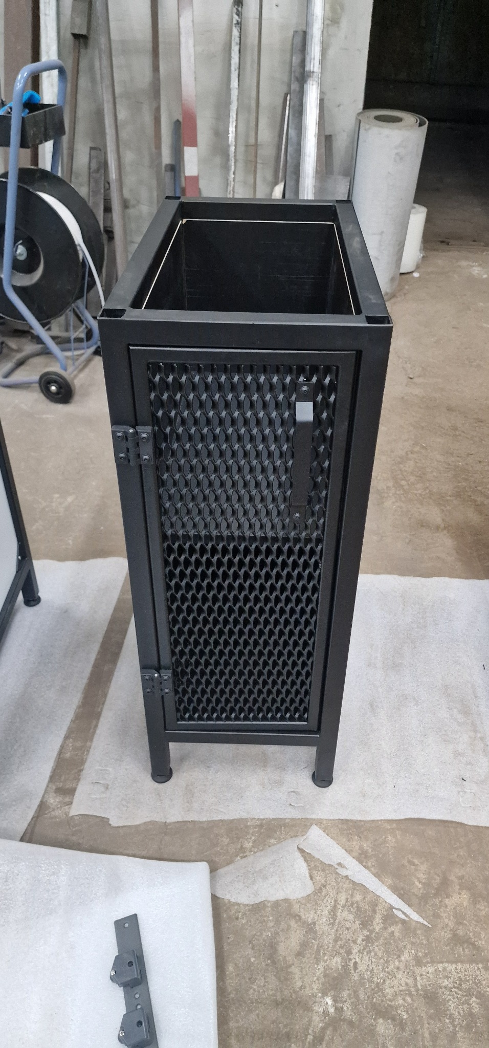 Czarna, metalowa szafka z perforowanymi drzwiczkami, widoczne zawiasy i uchwyt. W tle warsztat z elementami metalowymi. Nowoczesny design, surowy styl.