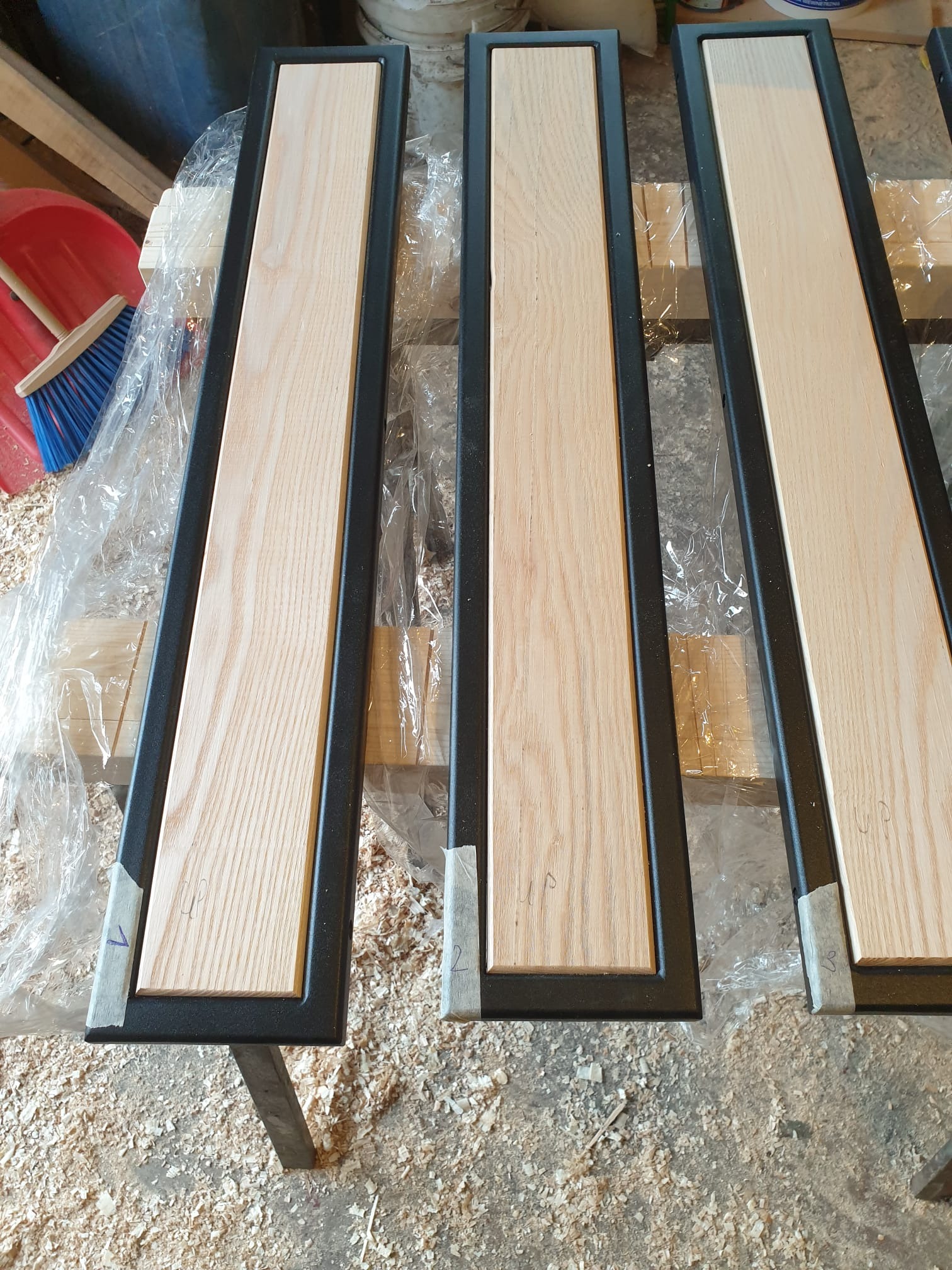 Elementy konstrukcyjne z drewna i metalu w warsztacie: trzy ramy z jasnym drewnem w czarnej metalowej obudowie, przygotowane do montażu, otoczone wiórami i narzędziami.