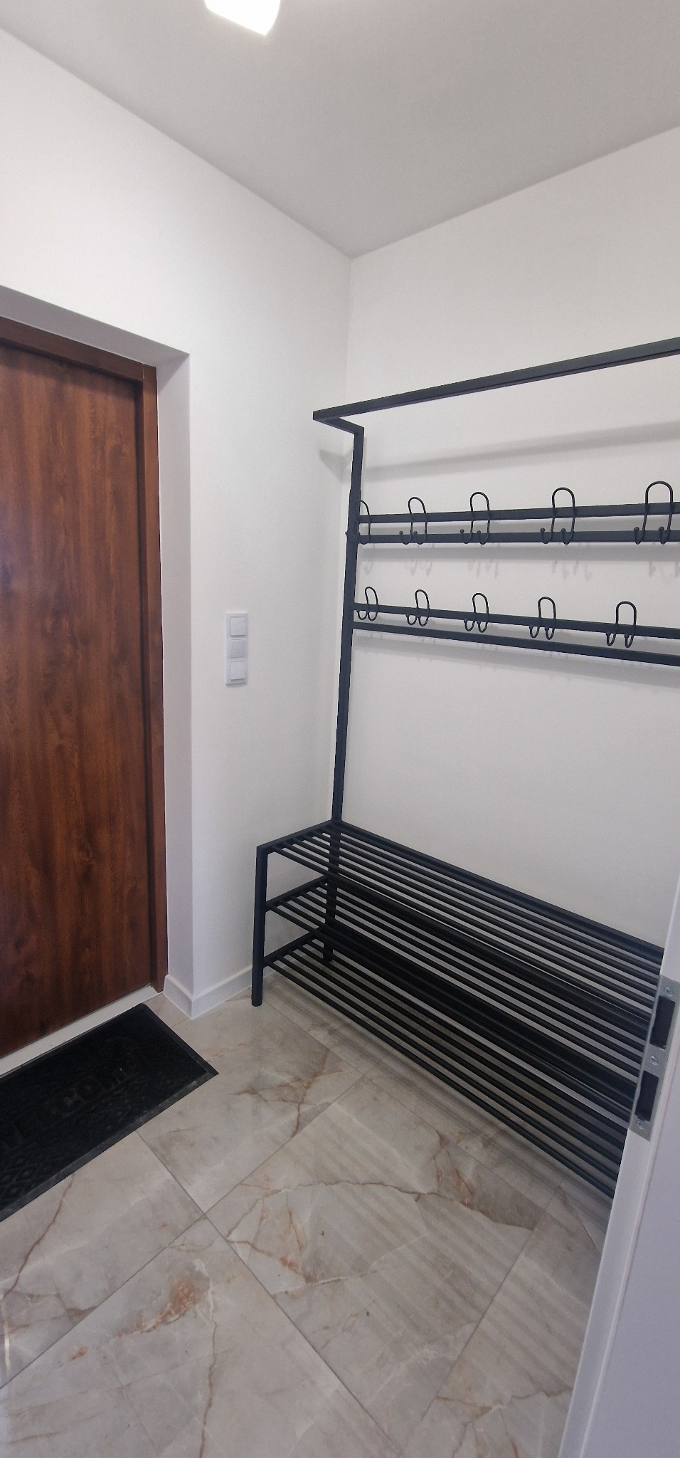 Nowoczesny, czarny metalowy wieszak na ubrania i buty w przedpokoju z brązowymi drzwiami, marmurową podłogą i białymi ścianami. Minimalistyczny design.