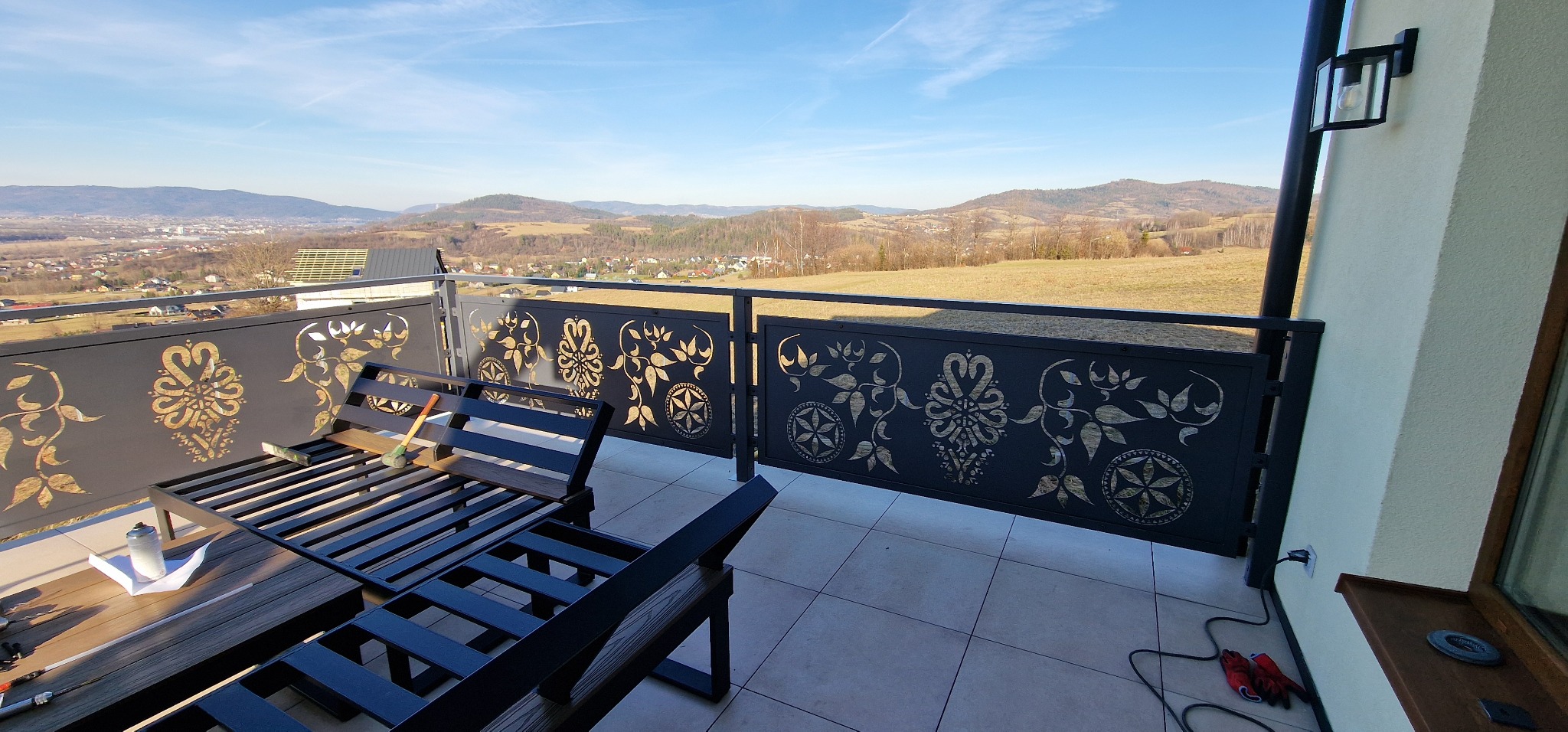 Czarna, metalowa balustrada balkonowa z ażurowym, złotym wzorem roślinnym, widok na górzysty krajobraz w tle. Na balkonie metalowe meble ogrodowe i narzędzia.