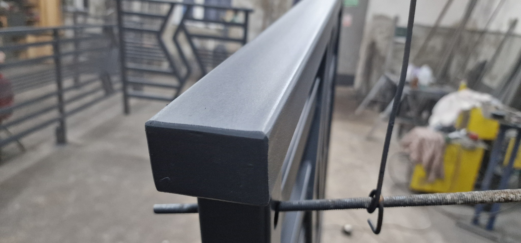 Szczegółowe ujęcie pomalowanej, metalowej balustrady z kwadratową poręczą. Widoczne ślady po zawieszeniu elementu podczas malowania. W tle warsztat.