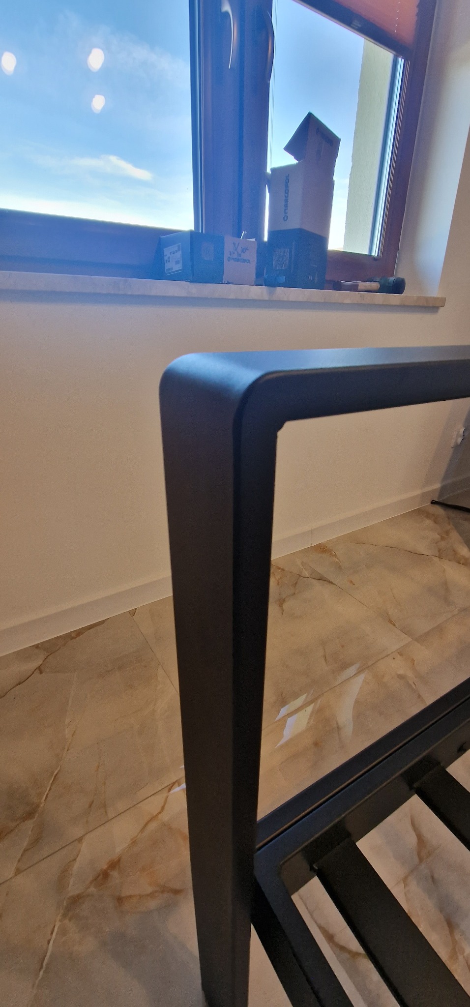 Fragment metalowej konstrukcji w ciemnym kolorze na tle okna. Widoczny marmurowy wzór na podłodze i elementy montażowe. Kąt ujęcia z dołu podkreśla solidność.