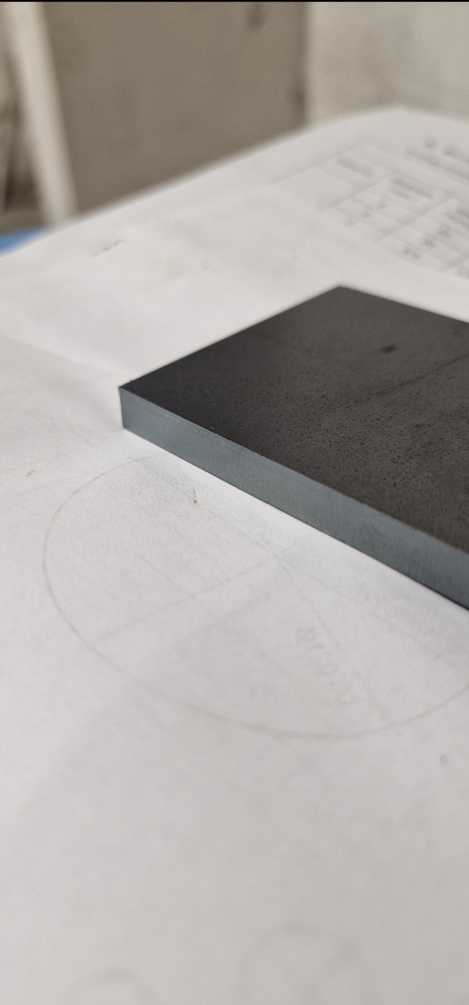 Precyzyjnie wycięty, ciemny element metalowy o gładkiej powierzchni, leżący na białym arkuszu z technicznym rysunkiem. Widoczny detal krawędzi i faktura materiału.