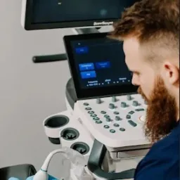 Lekarz w niebieskich rękawiczkach wykonuje USG kolana pacjenta, używając nowoczesnego aparatu ultrasonograficznego. Na ekranie widoczny obraz diagnostyczny.