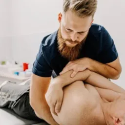 Mężczyzna z brodą wykonuje masaż barku pacjenta leżącego na kozetce w jasnym gabinecie. Widoczny fragment blatu z akcesoriami.