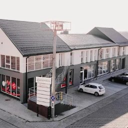 Barteczko Fizjoterapia Mysłowice - Rehabilitacja - Terapia Manualna - Osteopatia - Dwukondygnacyjny budynek centrum medycznego z szarym dachem, parkingiem i zielenią w tle. Tablica informacyjna przed wejściem. Widok z lotu ptaka.