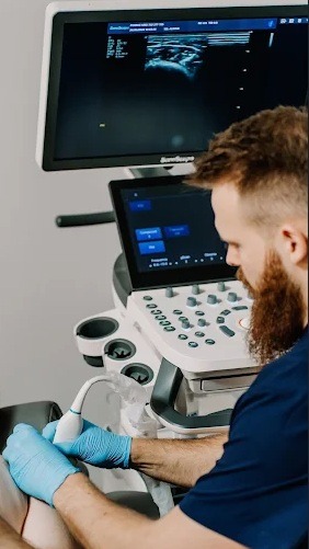 Lekarz w niebieskich rękawiczkach wykonuje USG kolana pacjenta, używając nowoczesnego aparatu ultrasonograficznego. Na ekranie widoczny obraz diagnostyczny.