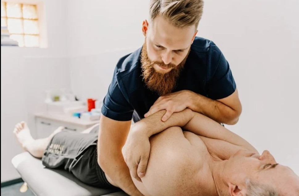 Mężczyzna z brodą wykonuje masaż barku pacjenta leżącego na kozetce w jasnym gabinecie. Widoczny fragment blatu z akcesoriami.