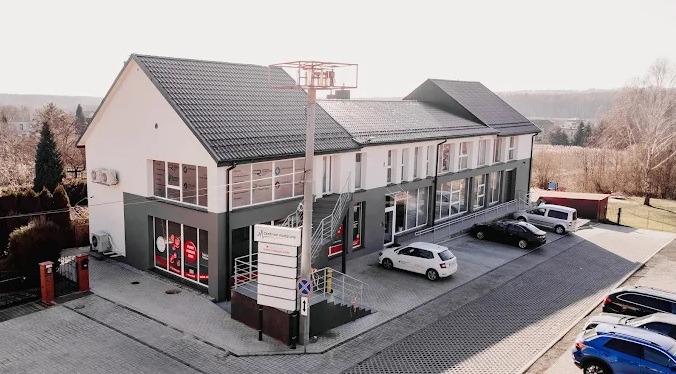 Dwukondygnacyjny budynek centrum medycznego z szarym dachem, parkingiem i zielenią w tle. Tablica informacyjna przed wejściem. Widok z lotu ptaka.