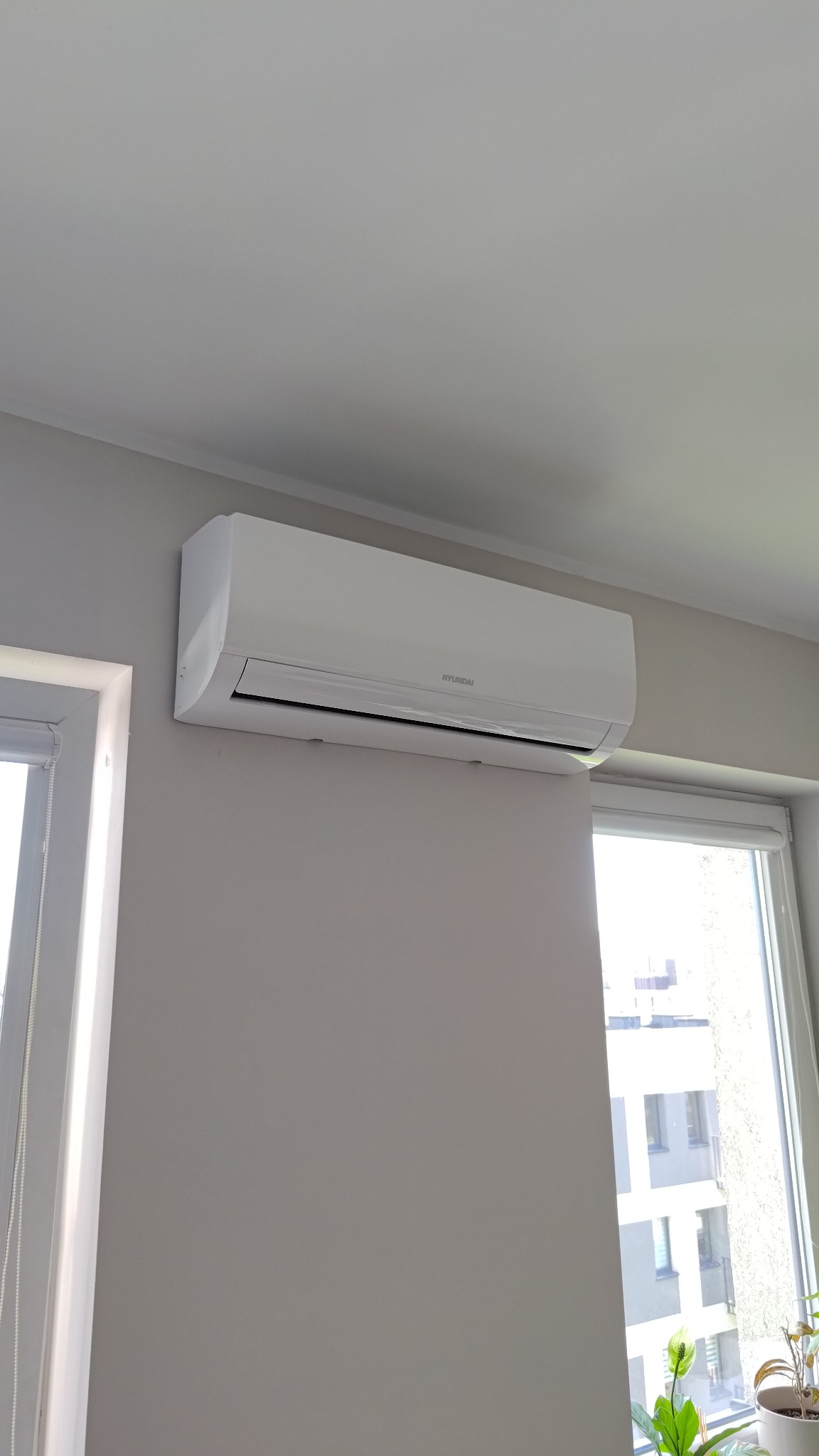 Biały klimatyzator Hyundai na szarej ścianie w jasnym wnętrzu, widok z dołu. Okno z roletą i roślina doniczkowa w tle.