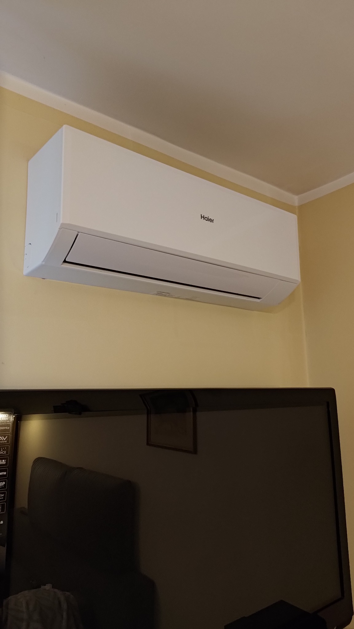 Biała klimatyzacja Haier, zamontowana na ścianie nad telewizorem w pokoju o beżowych ścianach. Widok z dołu, na urządzenie i fragment telewizora.