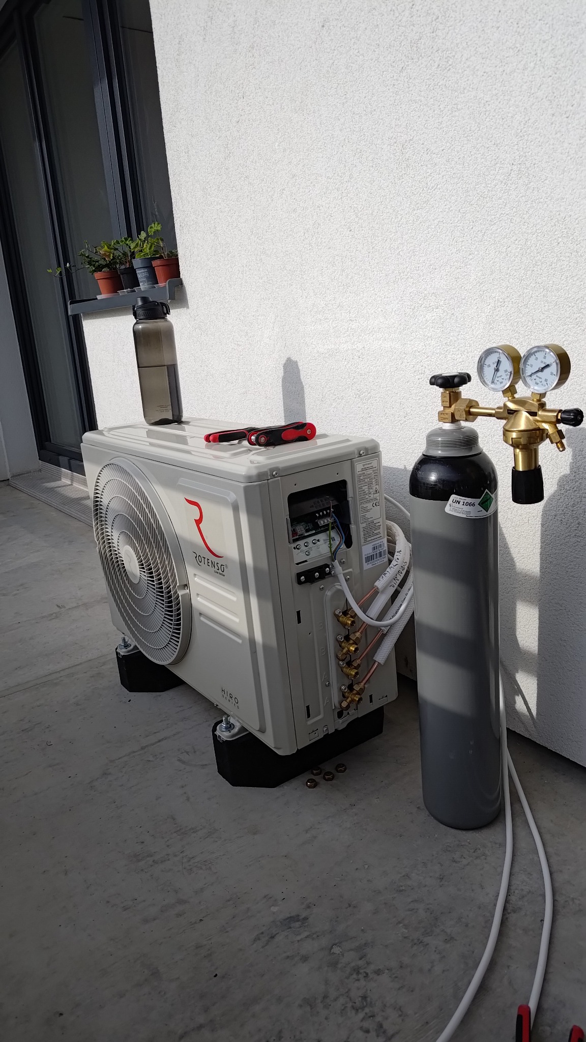 Biała jednostka zewnętrzna klimatyzacji Rotenso Hiro Series, butla z gazem i narzędzia na betonowym podłożu przy ścianie z oknem i doniczkami.