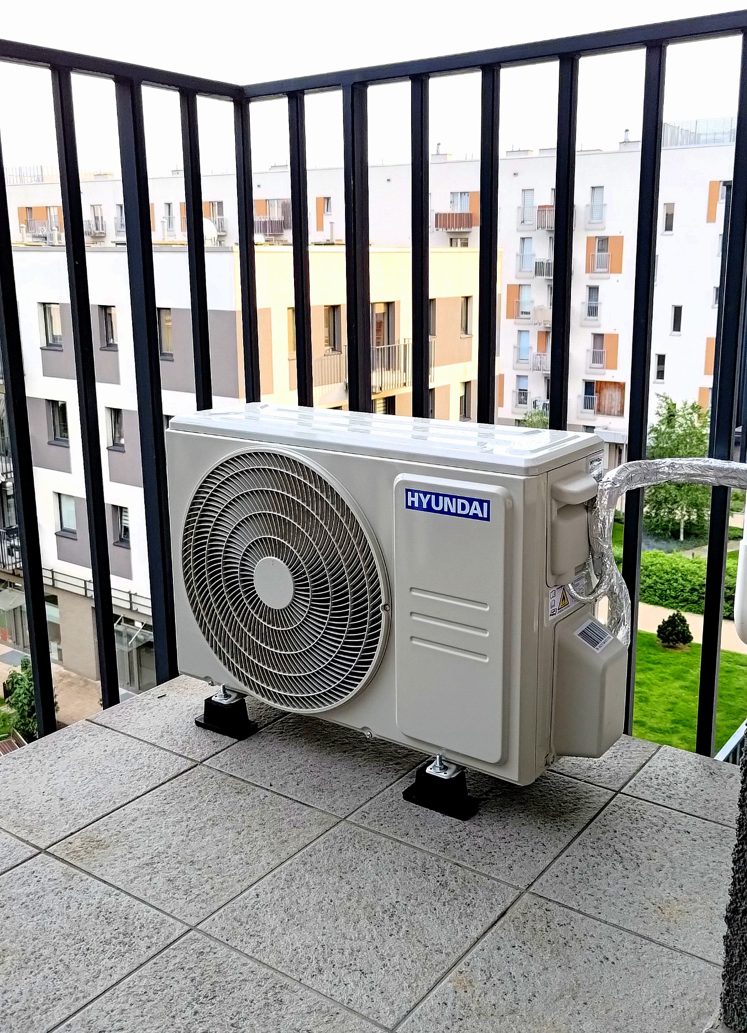 Zamontowana jednostka zewnętrzna klimatyzacji Hyundai na balkonie z widokiem na budynki mieszkalne i zieleń. Widoczne czarne podkładki pod urządzeniem.