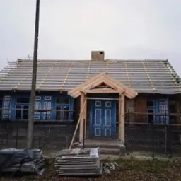 Drewniany dom z niebieską stolarką okienną i konstrukcją dachu w trakcie budowy. Widoczne elementy więźby dachowej i przygotowane materiały budowlane.