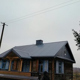 Grzegorz Litwiniuk DachStyl - Dom z ciemną, metalową blachodachówką i błękitnymi okiennicami, uchwycony w pochmurny dzień. Widoczne detale drewnianej konstrukcji i słup elektryczny w tle.