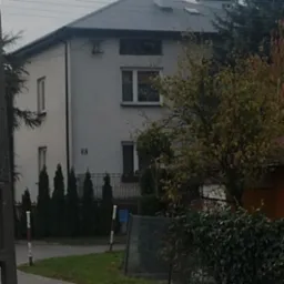 Dwupiętrowy dom jednorodzinny z szarym dachem i białą elewacją, otoczony zielenią. Widok z poziomu chodnika, drzewo po prawej stronie kadru. Ujęcie w pochmurny dzień.