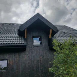 Ciemna elewacja domu z oknem i dachem pokrytym czarną dachówką, widok na fragment budynku i pochmurne niebo. Kontrast ciemnego drewna i jasnych refleksów w oknach.