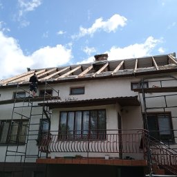 Grzegorz Litwiniuk DachStyl - Dwupiętrowy dom w trakcie wymiany dachu, widoczne rusztowania i pracownik na dachu. Częściowo zdjęte pokrycie, odsłonięta konstrukcja więźby dachowej. Błękitne niebo w tle.