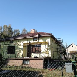 Grzegorz Litwiniuk DachStyl - Dom jednorodzinny w trakcie prac dekarskich, z rusztowaniem i pracownikiem na dachu. Widoczny fragment dachu z brązową dachówką i komin z cegły.