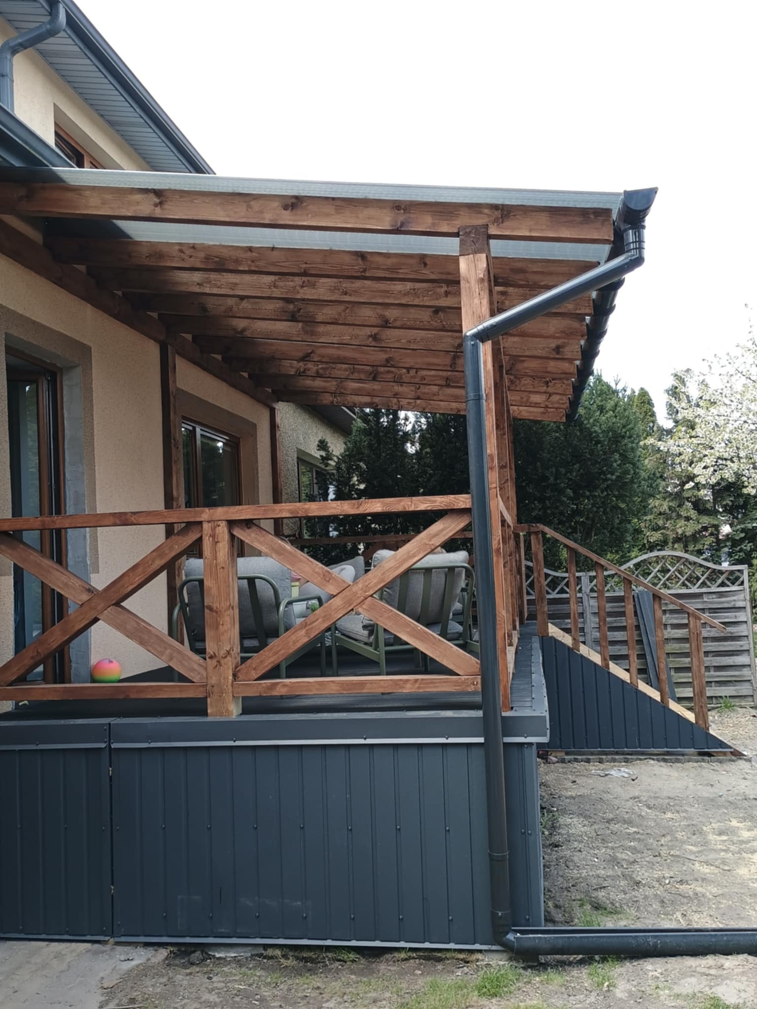 Drewniana pergola z rynnami przy domu, widok z boku. Konstrukcja z brązowego drewna, szare rynny. Na tarasie krzesła ogrodowe. W tle zieleń.