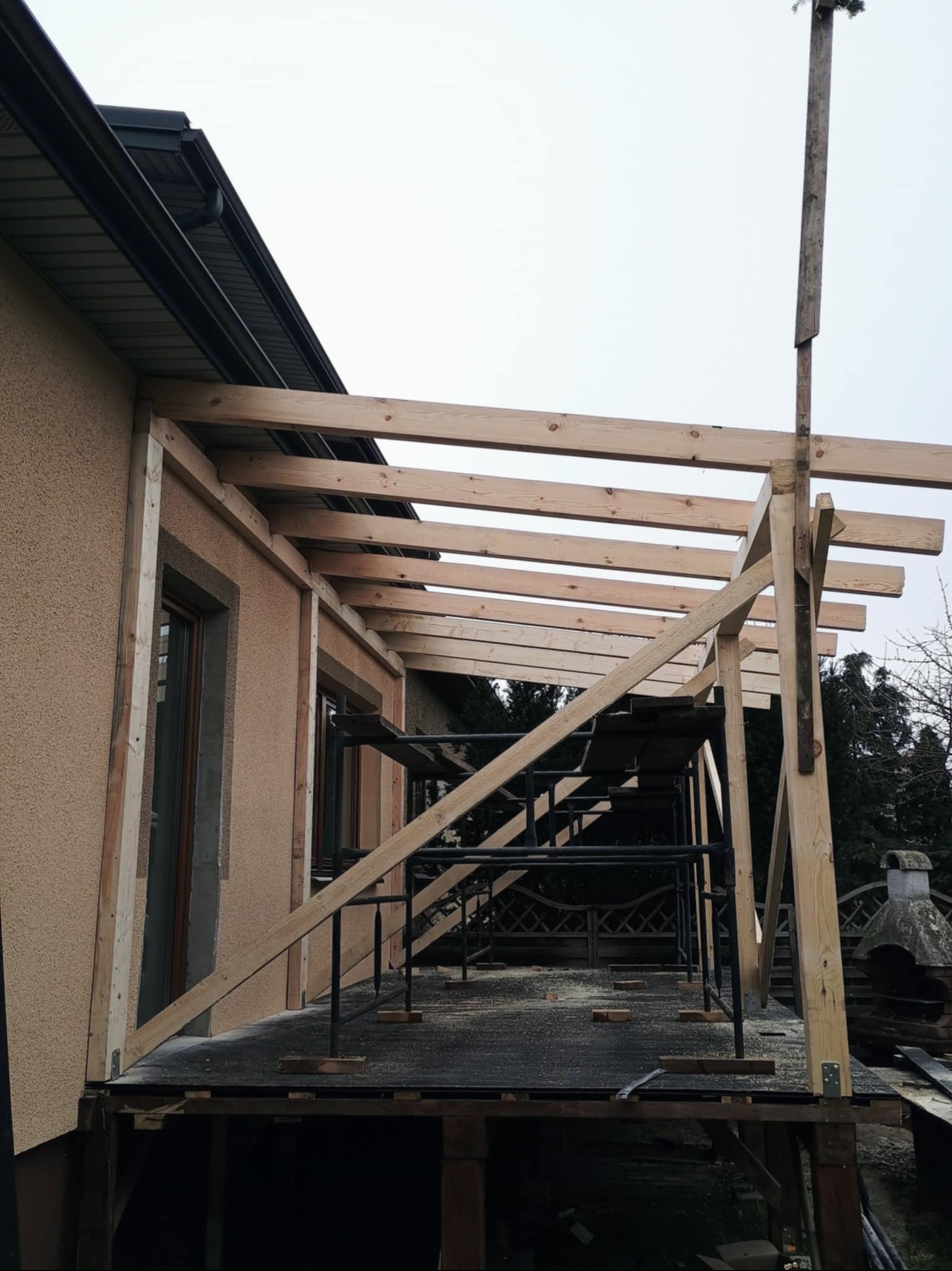 Drewniana konstrukcja z belek przy ścianie budynku, widoczne rusztowanie i fragment dachu z rynną. Budowa w toku, elementy montażowe, surowy stan wykończenia.