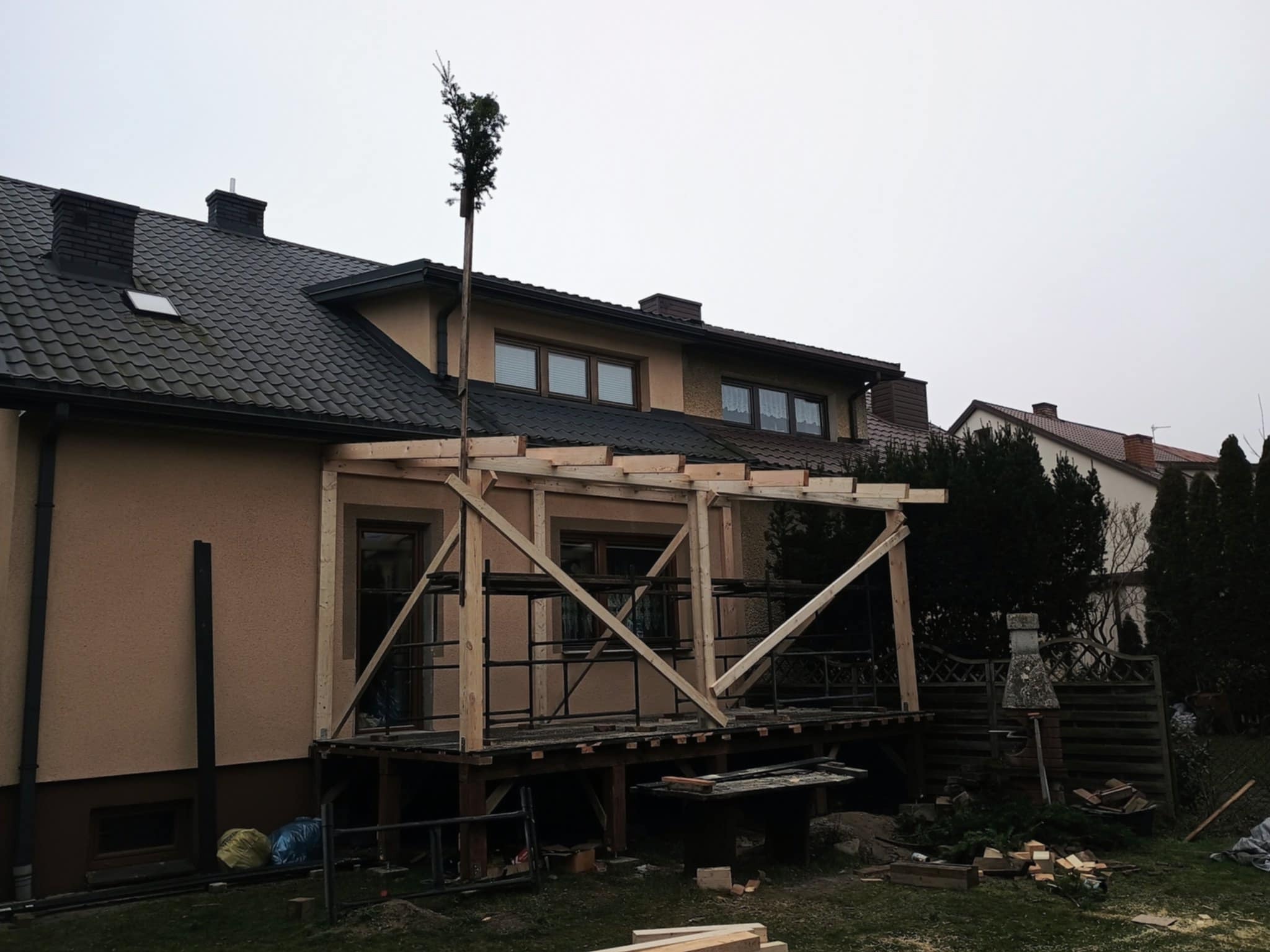 Budowa drewnianej konstrukcji zadaszenia tarasu przy domu z ciemnym dachem. Widoczne rusztowania i materiały budowlane. Etap w trakcie realizacji.