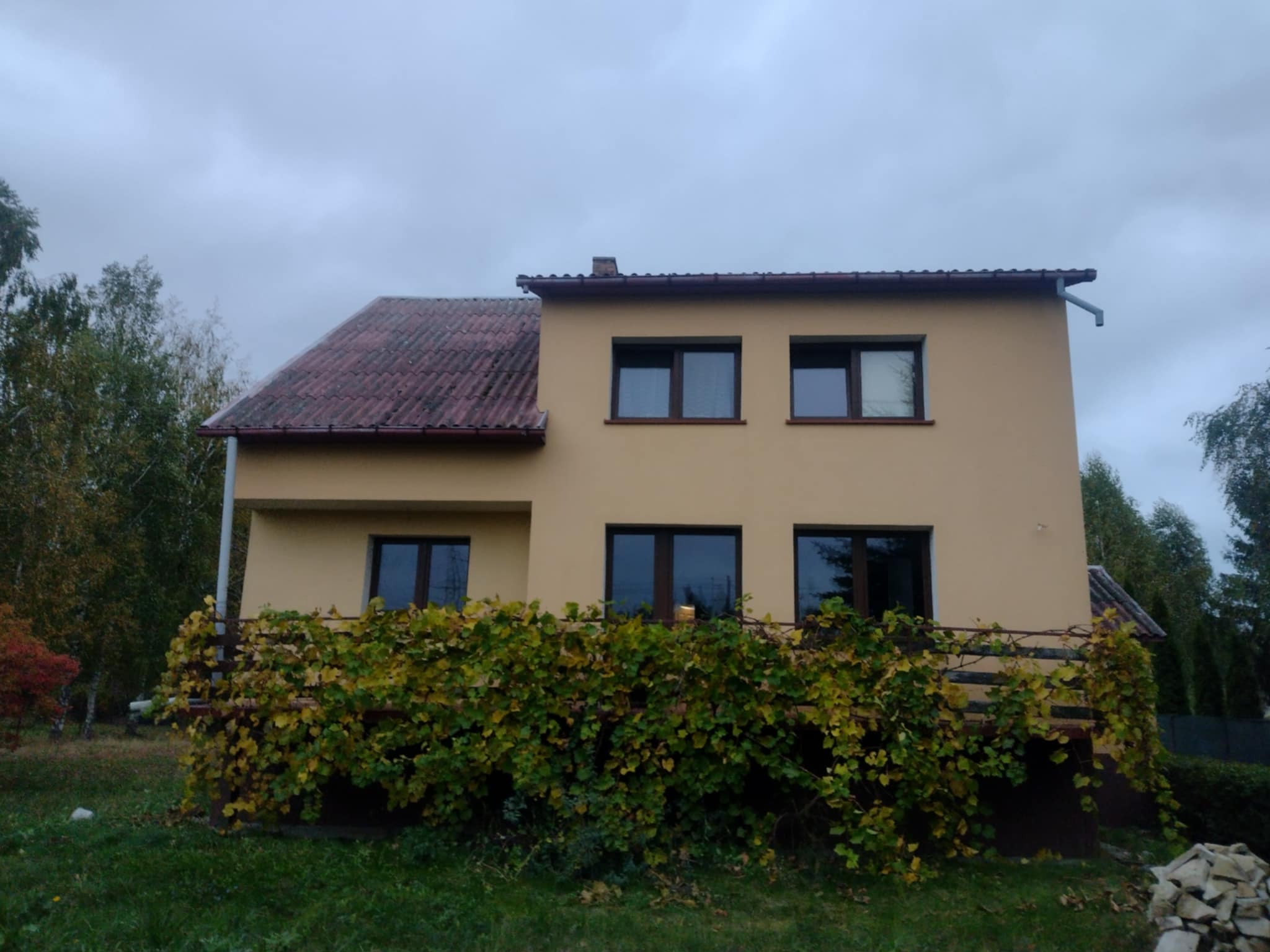 Dwupiętrowy dom z beżową elewacją i czerwoną dachówką, zarośnięty winoroślą na balkonie, sfotografowany w pochmurny dzień.