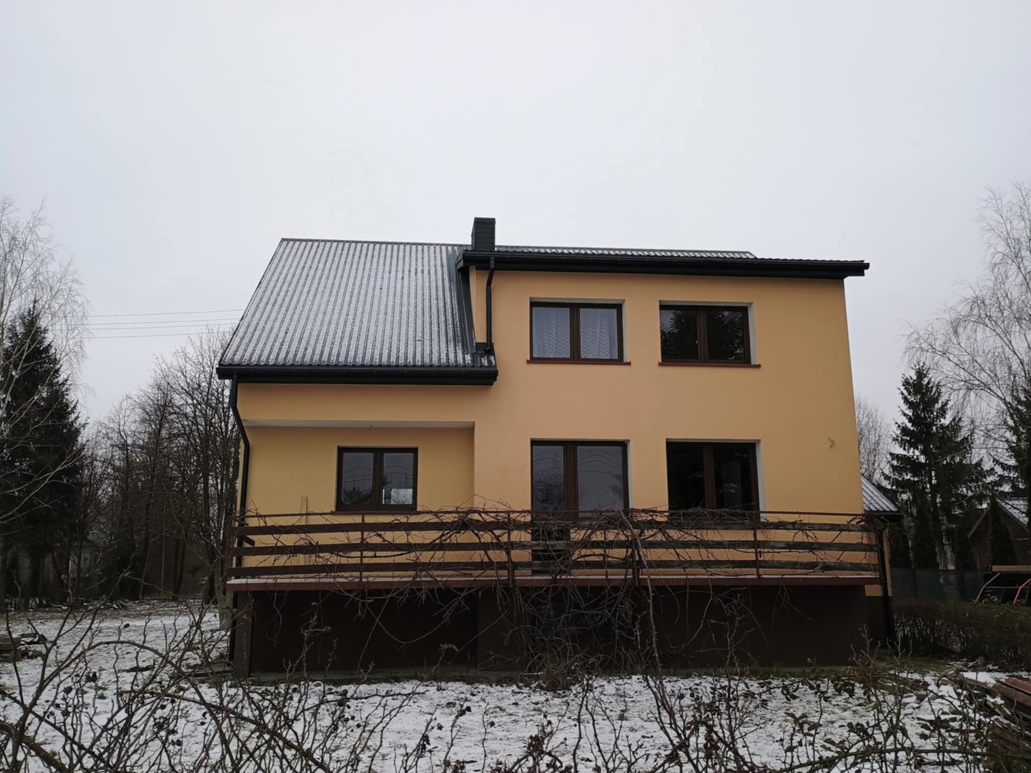 Dwukondygnacyjny dom z żółtą elewacją i szarym dachem pokrytym śniegiem, widziany w pochmurny dzień. Balustrada balkonu z drewnianych belek i gałęzi na pierwszym planie.