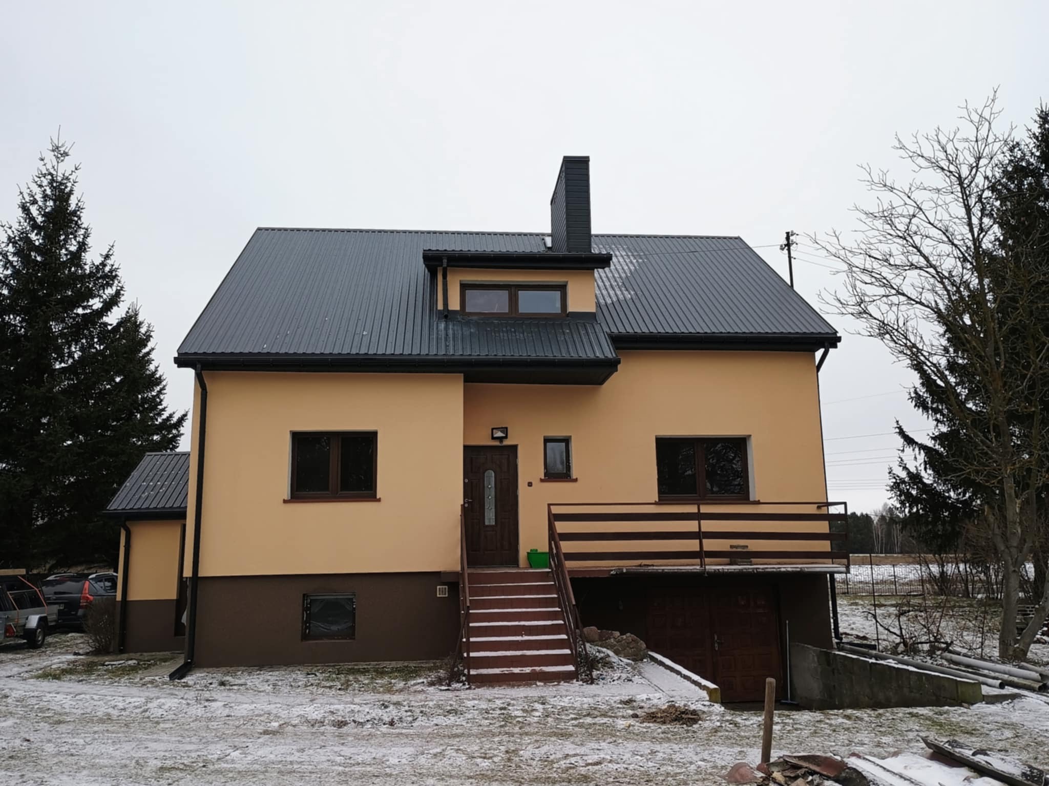 Dom jednorodzinny z nowym, ciemnym dachem z blachy trapezowej i ocieploną elewacją w odcieniach beżu i brązu, z elementami wykończenia w kolorze ciemnego drewna.