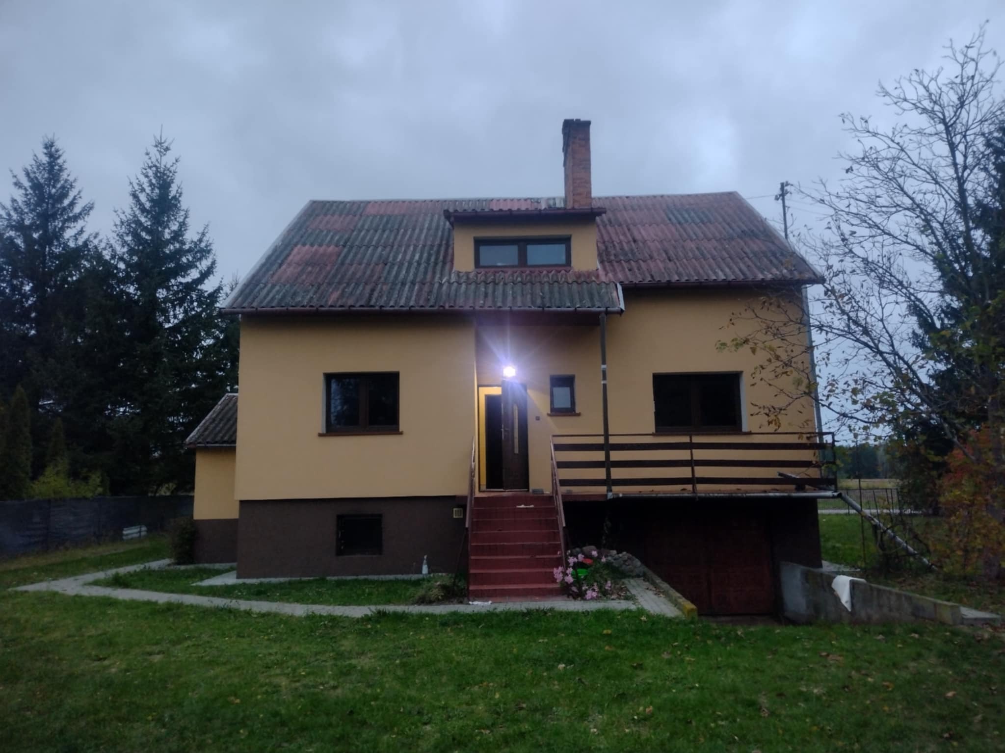 Dwukondygnacyjny dom z czerwonymi schodami wejściowymi i dachem z szarej dachówki, otoczony zielonym trawnikiem i drzewami pod pochmurnym niebem.