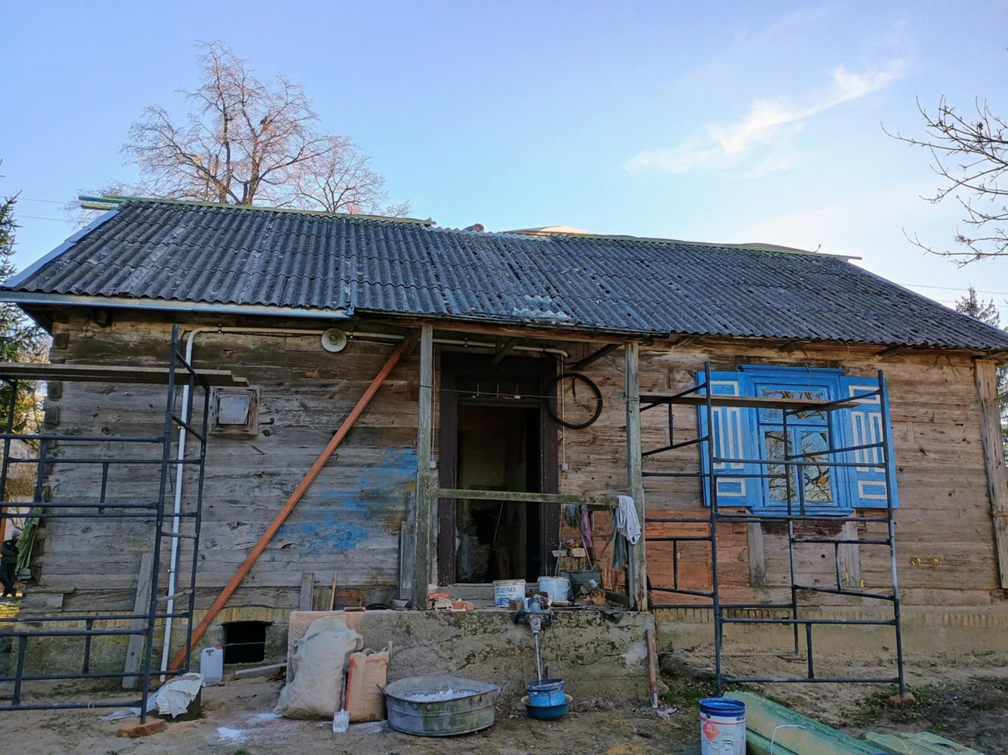 Drewniany dom w trakcie renowacji: widoczne rusztowania, worki z zaprawą, mieszadło i remontowane okno z niebieskimi okiennicami. Dach z eternitu.