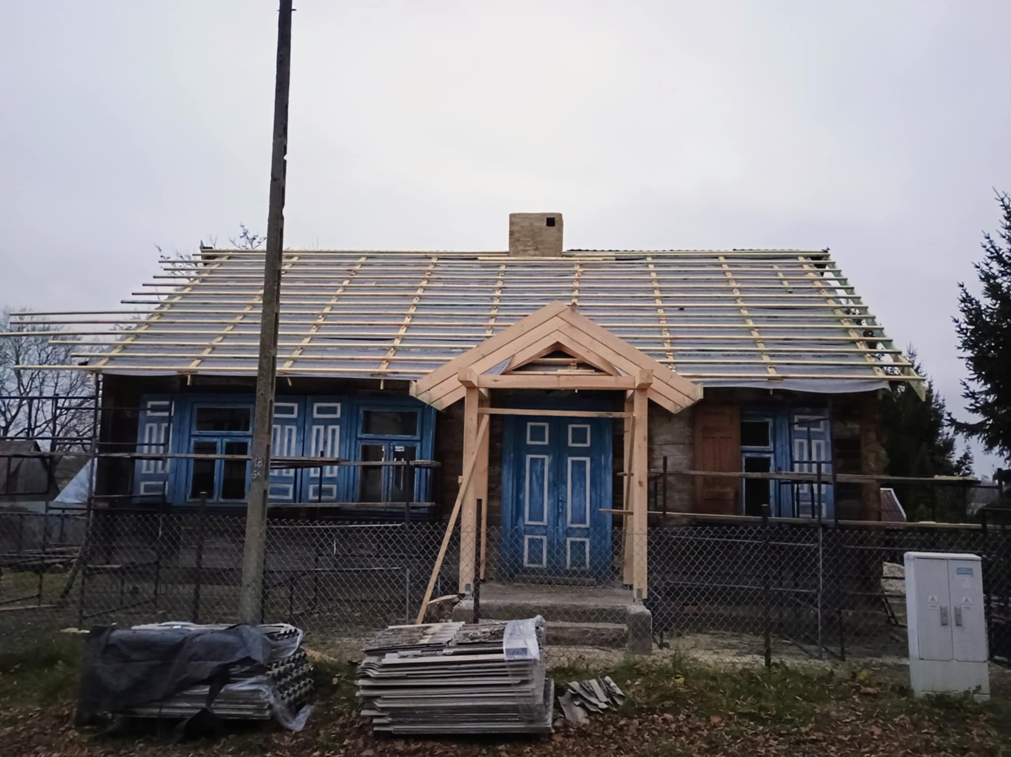 Drewniany dom z niebieską stolarką okienną i konstrukcją dachu w trakcie budowy. Widoczne elementy więźby dachowej i przygotowane materiały budowlane.