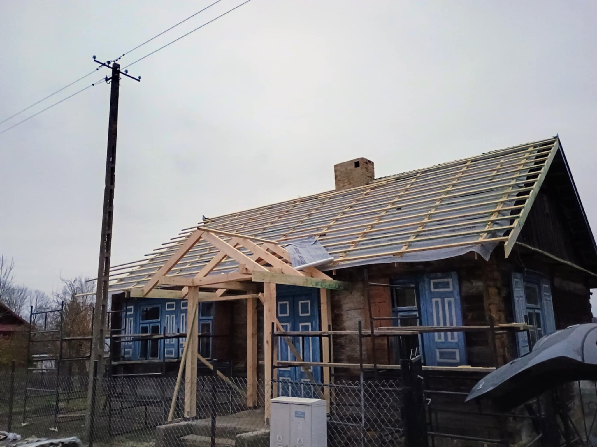 Drewniany dom w trakcie budowy ganku i wymiany pokrycia dachowego. Widoczne krokwie i łaty na dachu, konstrukcja ganku z surowego drewna, błękitne okiennice.