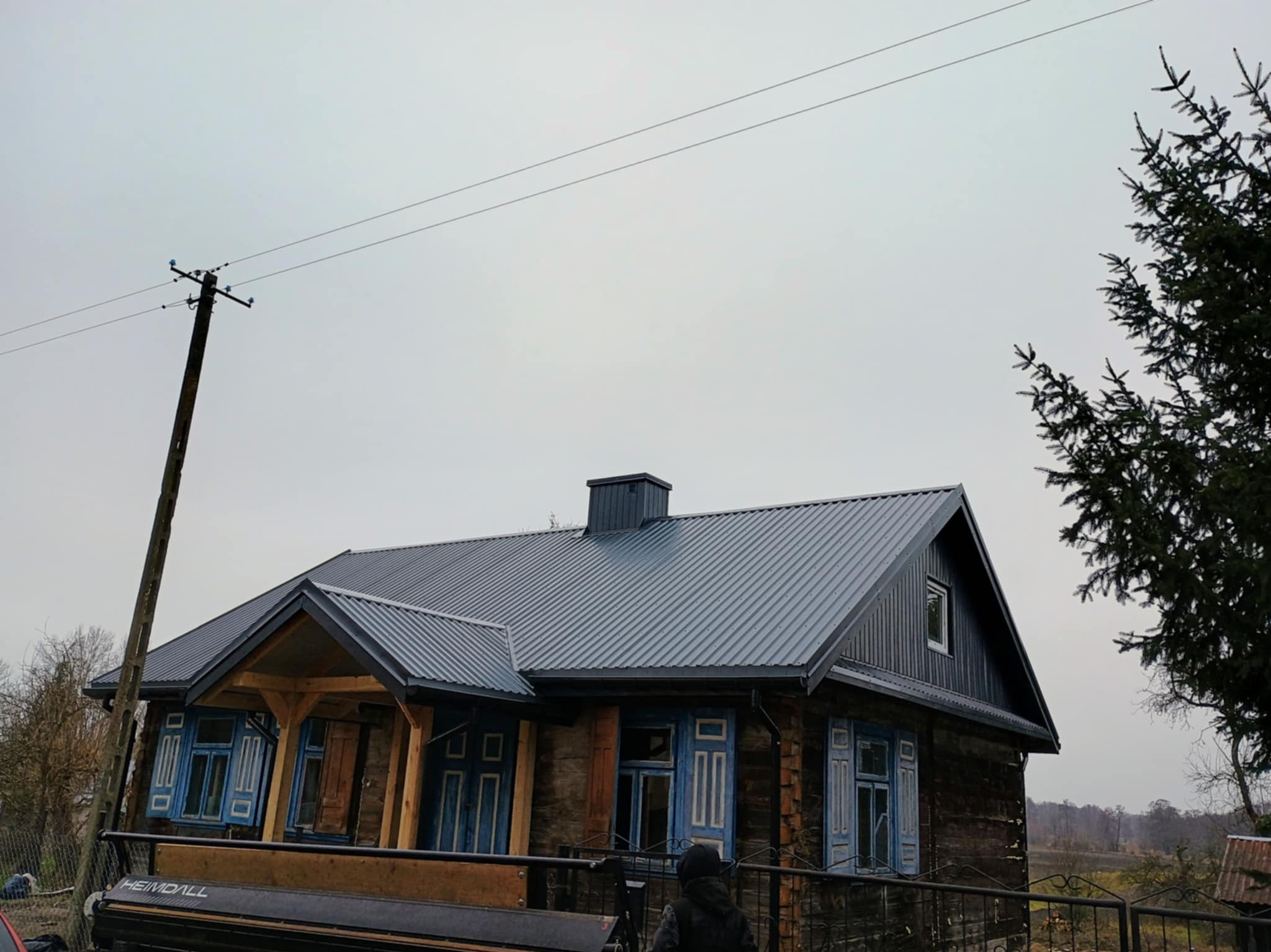 Dom z ciemną, metalową blachodachówką i błękitnymi okiennicami, uchwycony w pochmurny dzień. Widoczne detale drewnianej konstrukcji i słup elektryczny w tle.