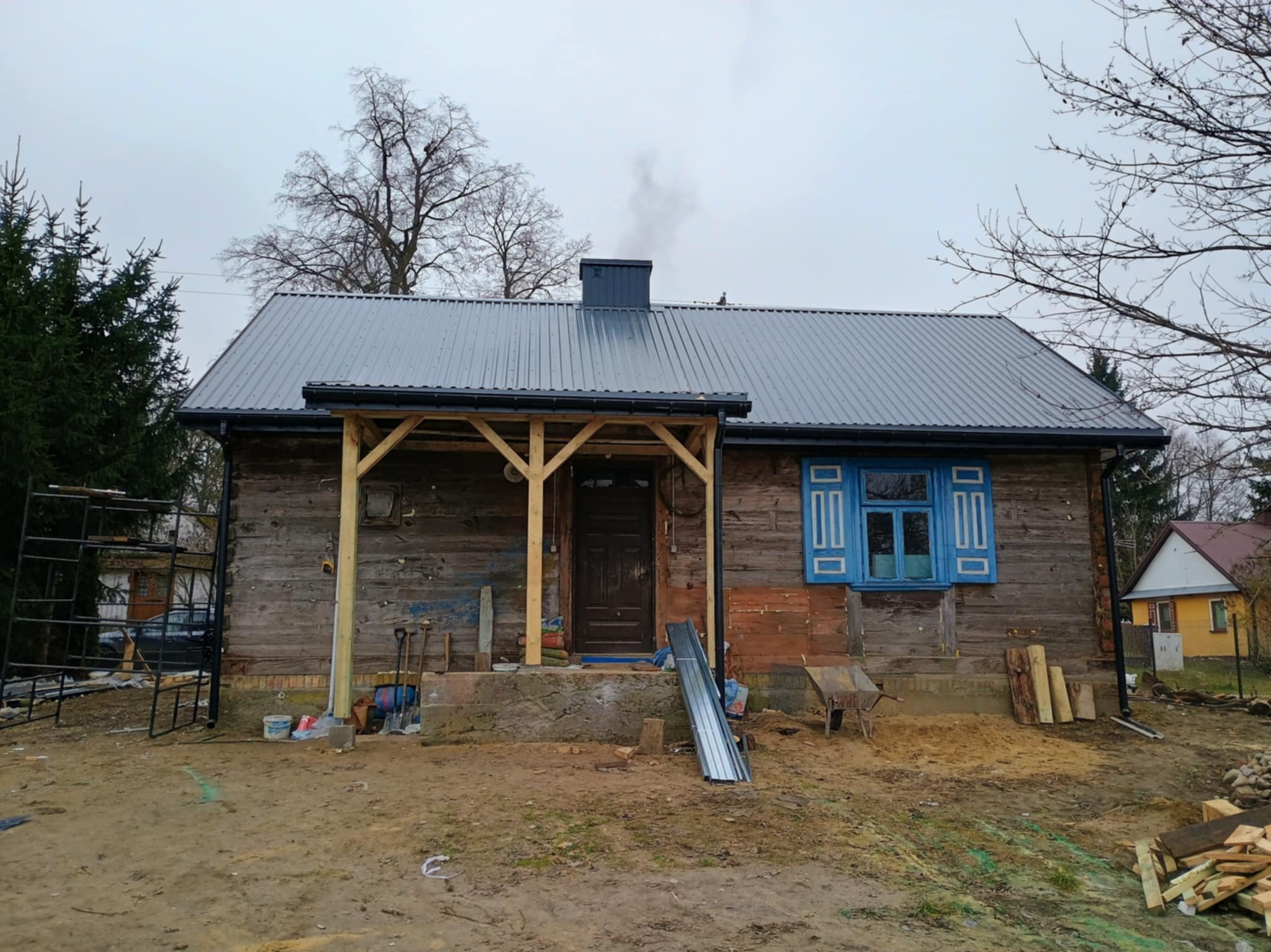 Wiejski dom z nowym, szarym dachem z blachy trapezowej i drewnianą werandą w trakcie budowy. Widoczne materiały budowlane i narzędzia. Niebieskie okiennice dodają koloru.