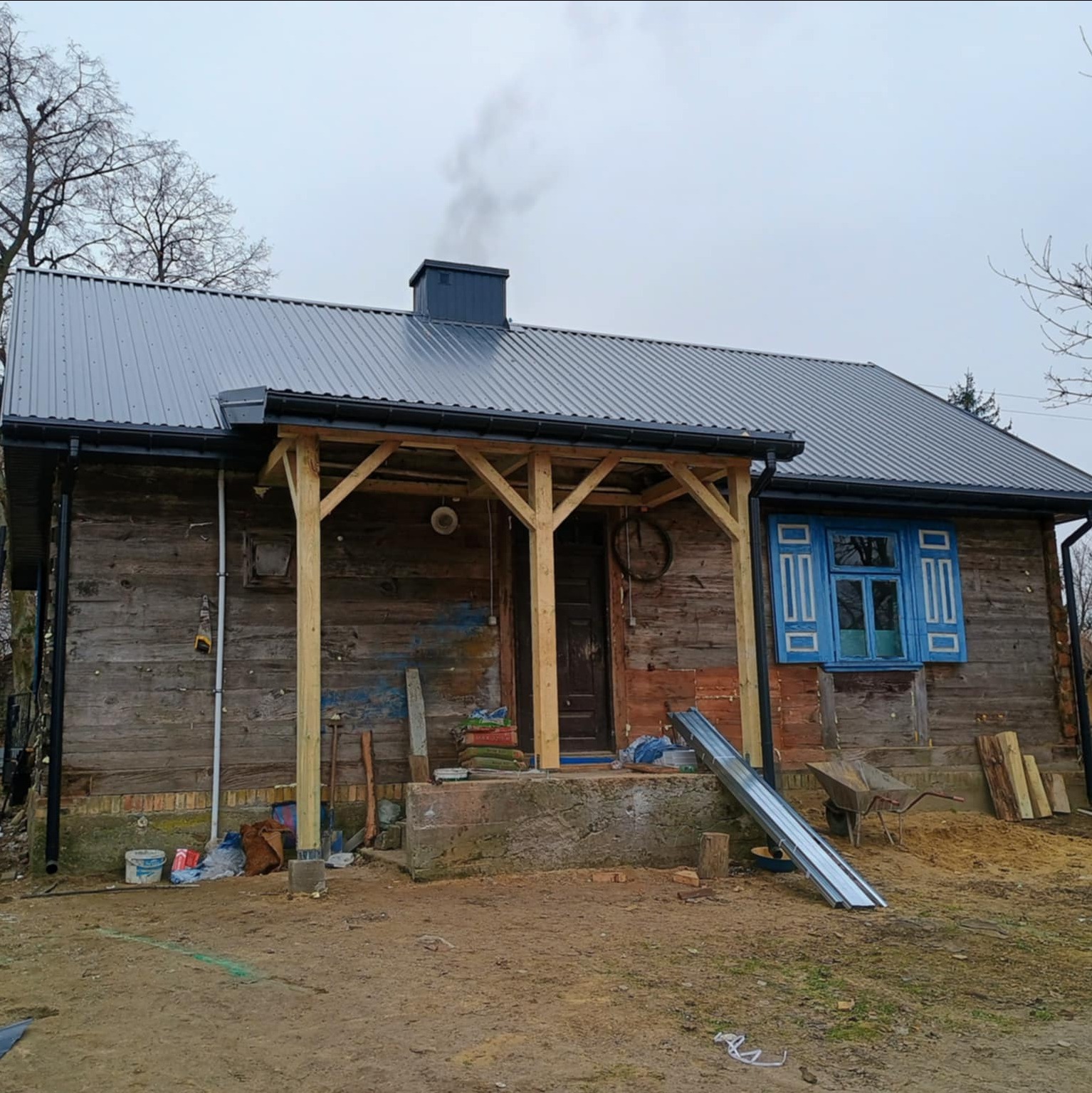 Dom z nowym, szarym dachem z blachy trapezowej i drewnianą werandą w trakcie budowy. Widoczne narzędzia i materiały budowlane.