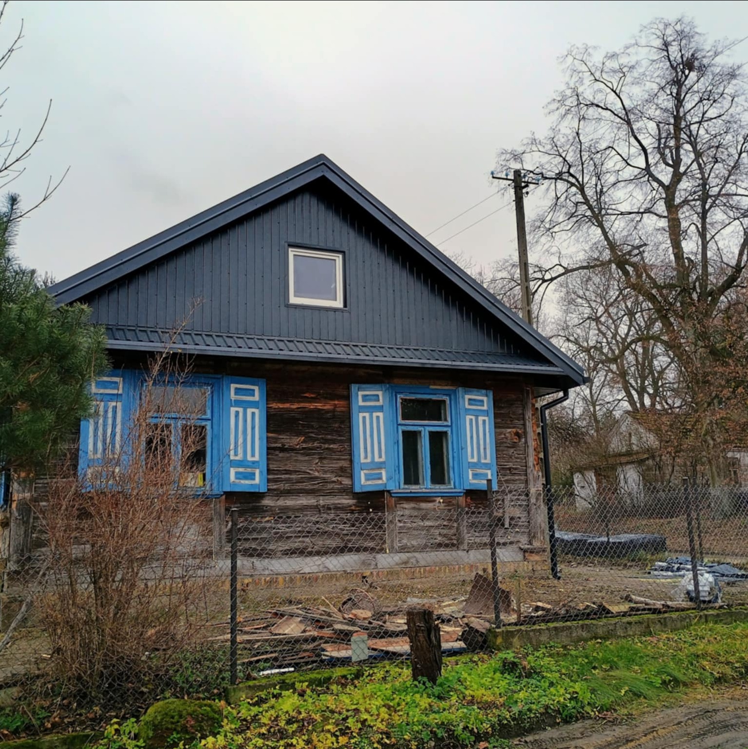 Drewniany dom z nowym, ciemnym dachem i niebieskimi okiennicami. Widoczne ślady prac remontowych wokół budynku i jesienne otoczenie.