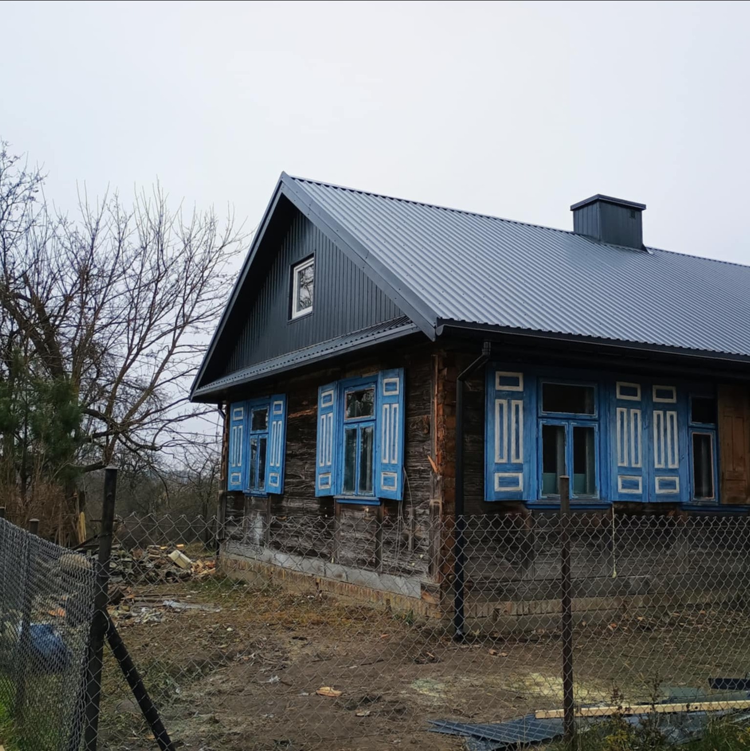 Dom z ciemnoszarym dachem z blachy trapezowej i niebieskimi okiennicami. Widok z zewnątrz, częściowo ogrodzony siatką.