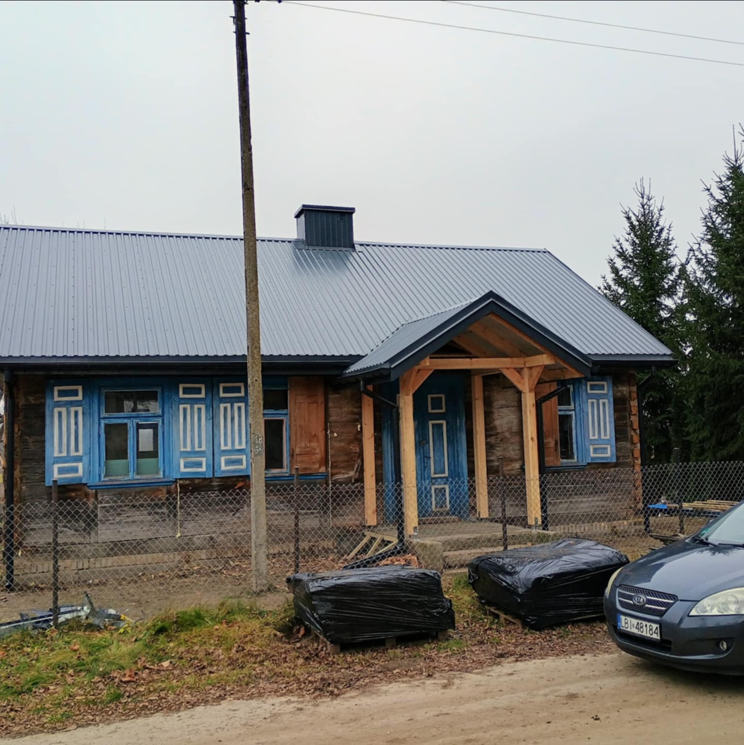 Dom z nowym, szarym dachem z blachy trapezowej, drewnianą werandą i niebieskimi okiennicami. Częściowo widoczny samochód i materiały budowlane w folii.