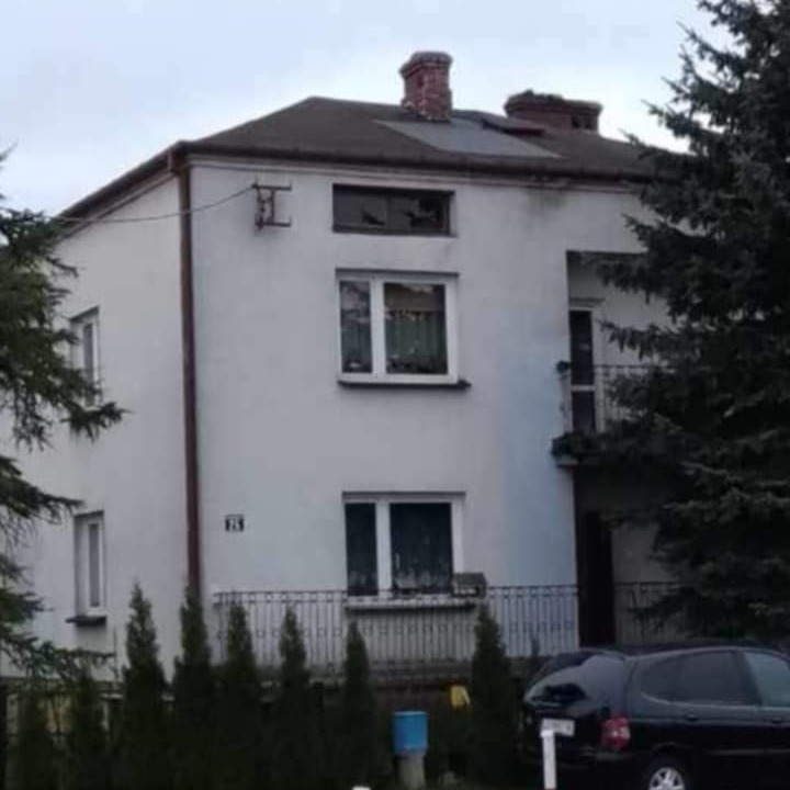 Dwupiętrowy dom z białymi ścianami i ciemnym dachem, widoczny z zewnątrz, z fragmentem czarnego samochodu na pierwszym planie i zielenią po bokach.