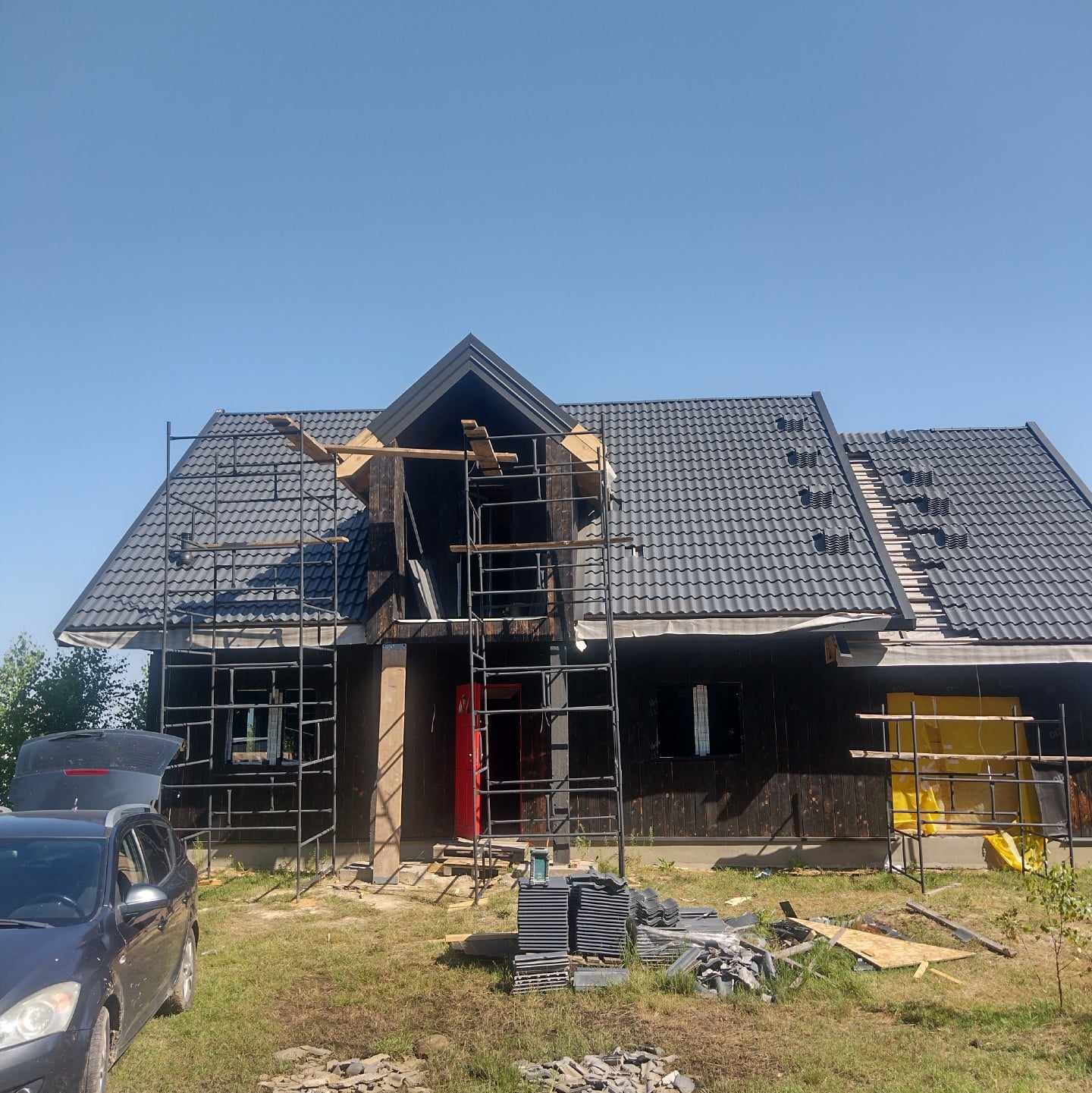 Dom w trakcie krycia dachu szarą dachówką, widoczne rusztowania i materiały budowlane. Część dachu już pokryta, reszta w trakcie prac. Słoneczny dzień.