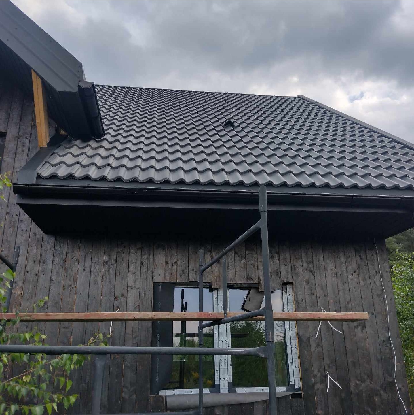 Czarny dach z blachy trapezowej na domu z ciemnego drewna, widok z rusztowania. Nowe okna w trakcie montażu, pochmurne niebo w tle.