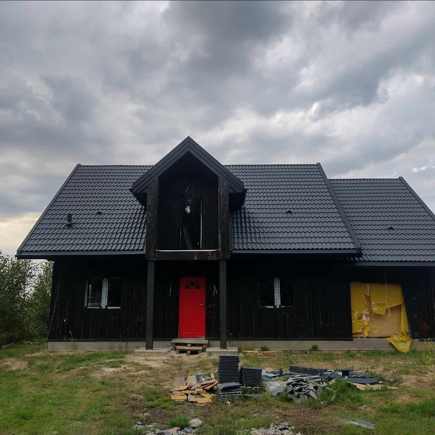 Dom z ciemną elewacją i nowym dachem z dachówki w Białej Podlaskiej. Czerwone drzwi wejściowe, widoczne materiały budowlane. Pochmurne niebo w tle.