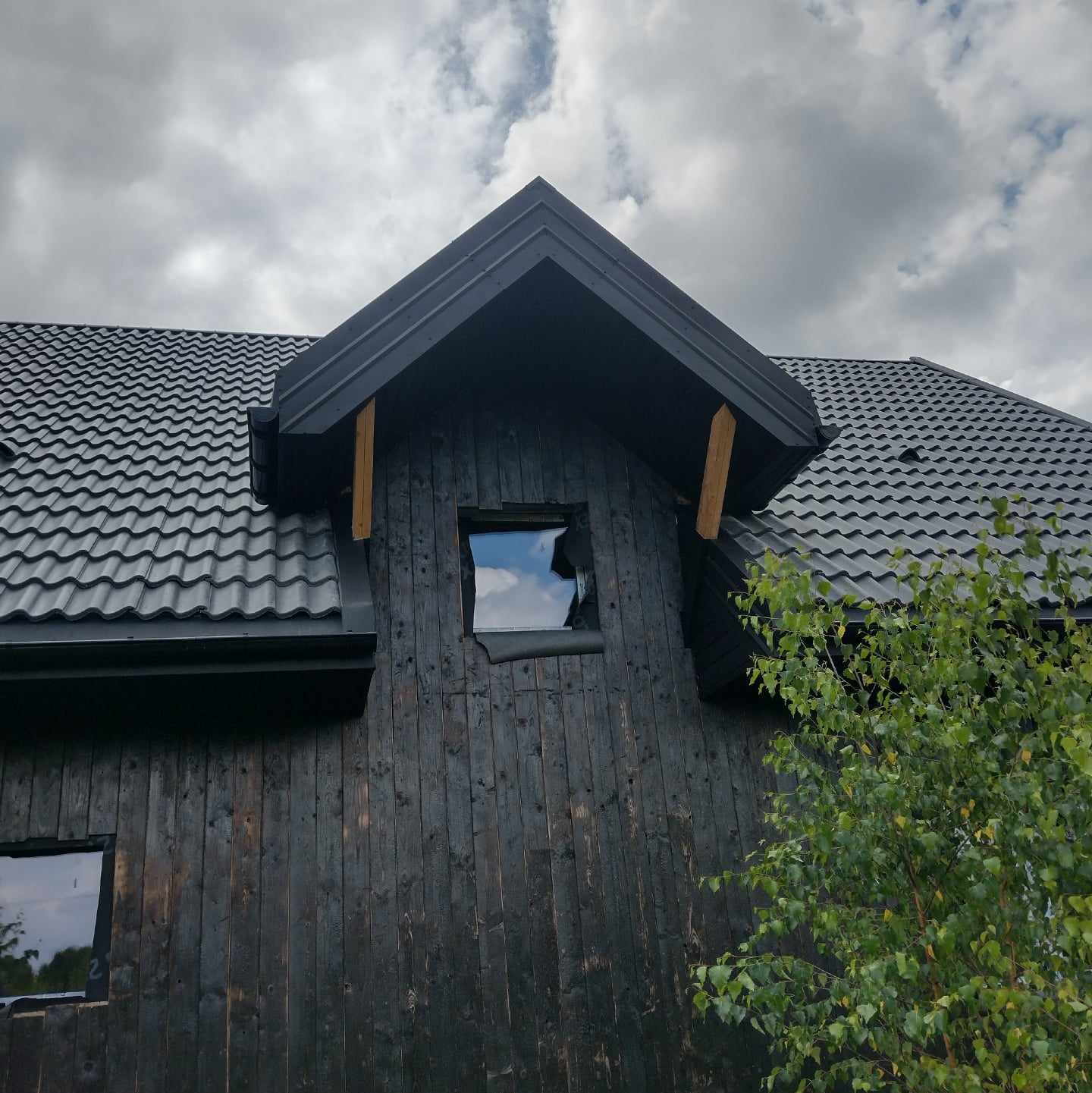 Ciemna elewacja domu z oknem i dachem pokrytym czarną dachówką, widok na fragment budynku i pochmurne niebo. Kontrast ciemnego drewna i jasnych refleksów w oknach.