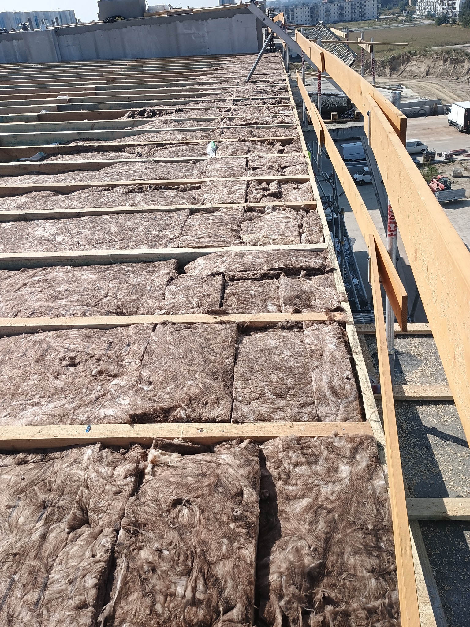 Ocieplenie dachu wełną mineralną, widok z góry na konstrukcję drewnianą i izolację termiczną. Prace dekarskie w toku, budowa dachu z widocznym ociepleniem.