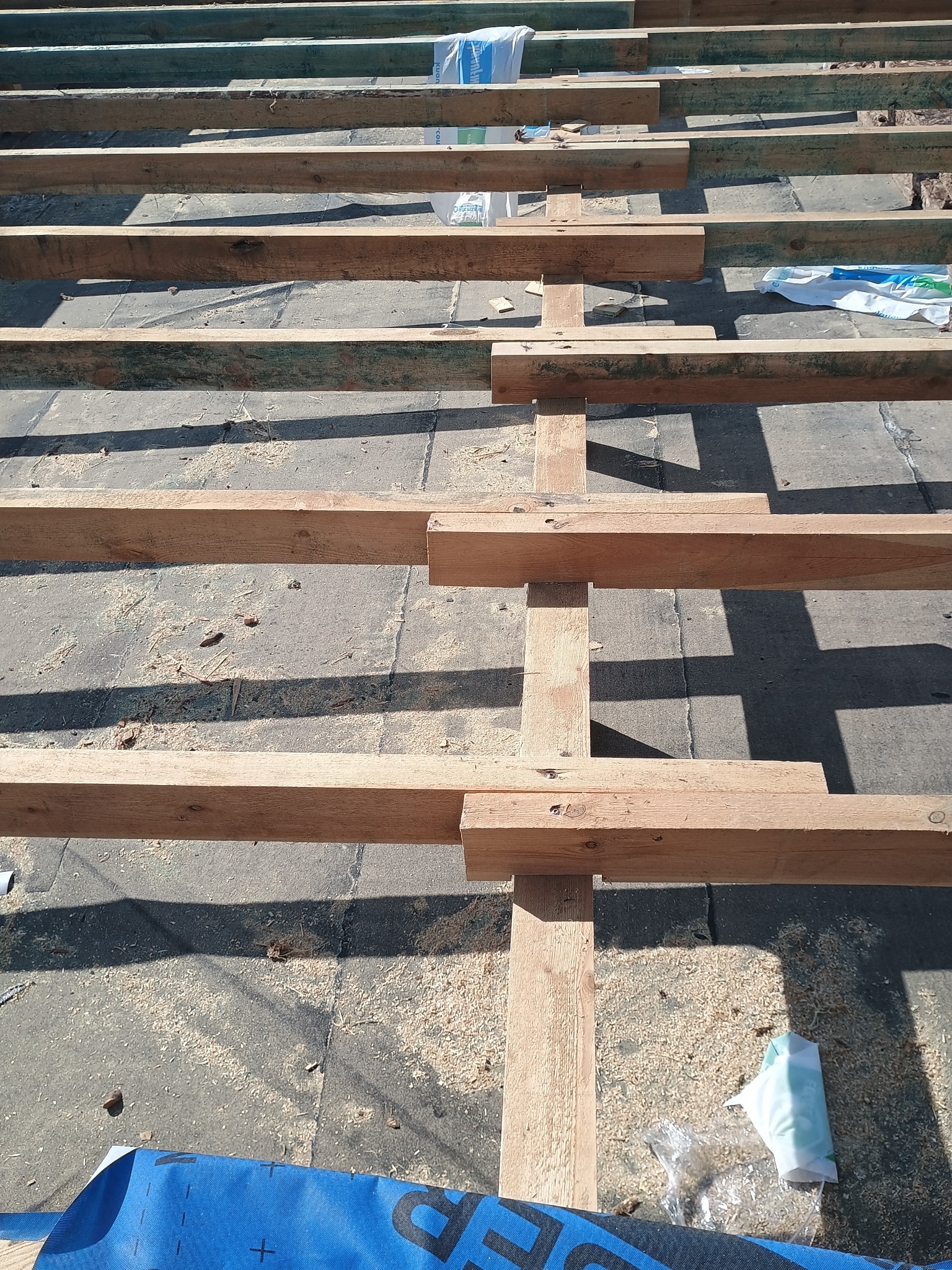 Konstrukcja drewniana dachu w trakcie budowy, widok z góry. Elementy drewniane ułożone równolegle, z widocznymi śladami obróbki i fragmentami materiałów budowlanych.