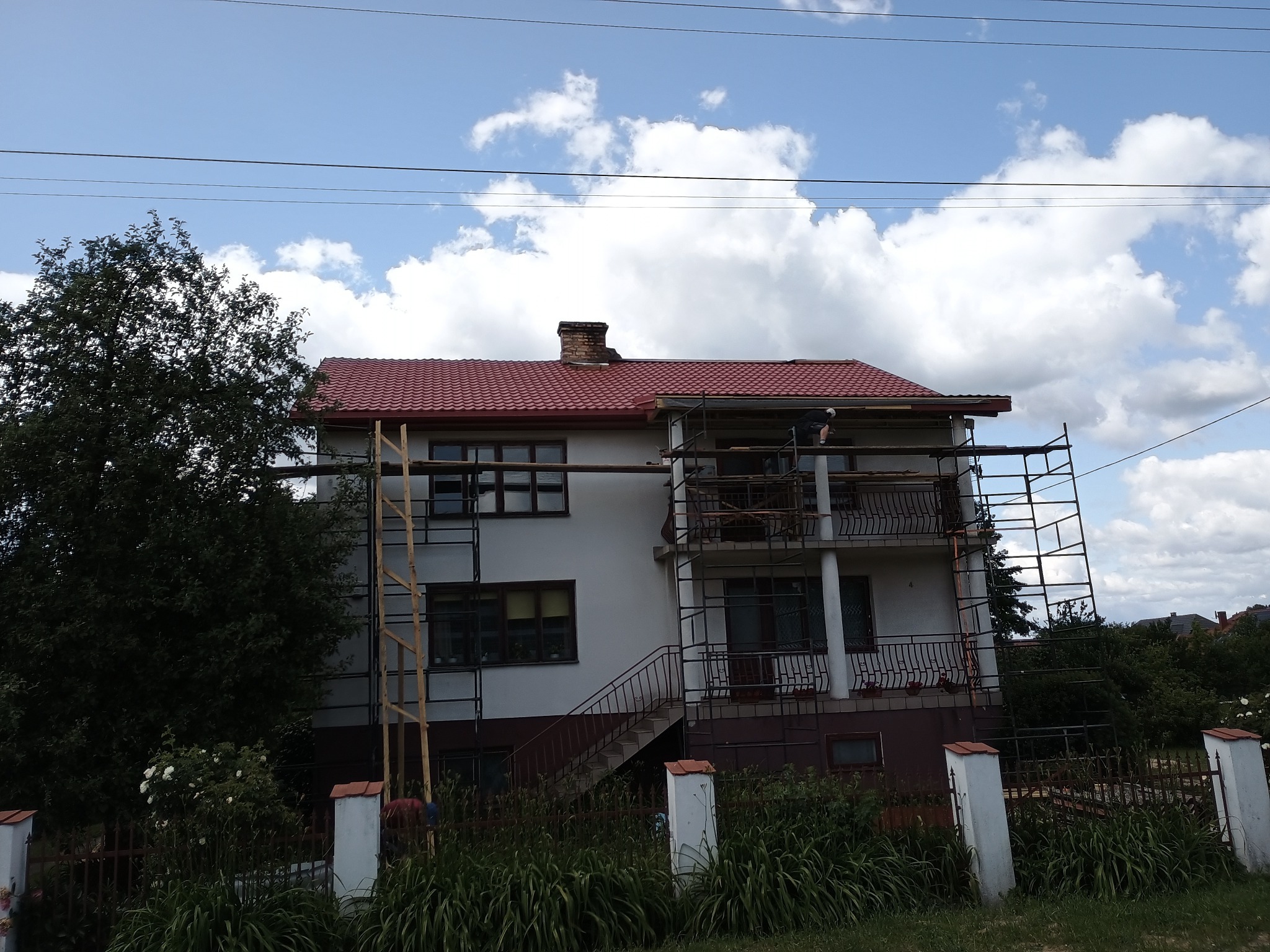 Dom jednorodzinny z nowym, czerwonym dachem z blachodachówki, otoczony rusztowaniami. Widoczny fragment komina i błękitne niebo z chmurami w tle.